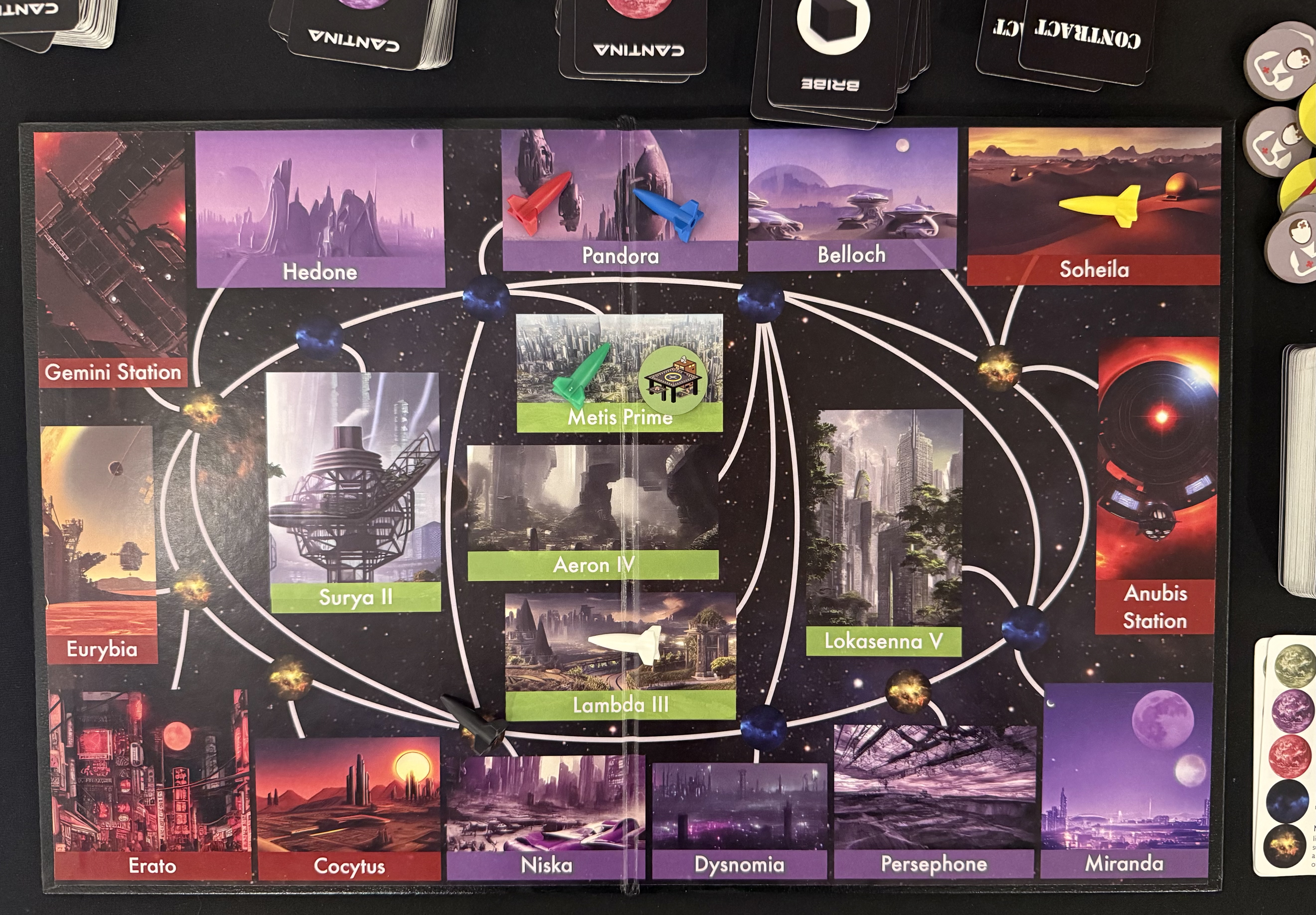A photo of the S.P.A.C.E. game board at the start of the sample turn showing the following planets and space stations: Hedone, Pandora, Belloch, Soheila, Gemini Station, Eurybia, Erato, Cocytus, Niska, Dysnomia, Persephone, Miranda, Surya II, Aeron IV, Lambda III, Metis Prime, Lokasenna V, and Anubis Station. Six colored ship tokens — blue, red, yellow, green, white, and black — are placed on various planets. Several decks of cards (Cantina and Contract) and stacks of small circular tokens are visible around the board edges. The map shows white connecting lines between locations representing possible travel routes, with art depicting space and alien worlds.